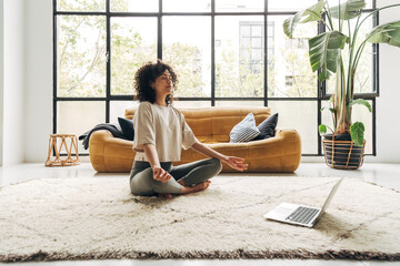 Young multiracial latina woman meditating at home with online video meditation lesson using laptop. - Powered by Adobe