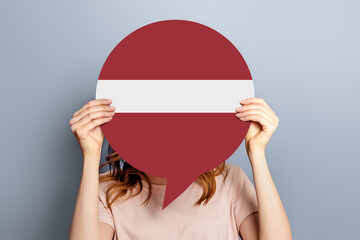 Voting in latvia concept. Student girl holds blank white speech bubble with latvia flag isolated over grey background. woman holding a round information piece of paper dialog - obrazy, fototapety, plakaty