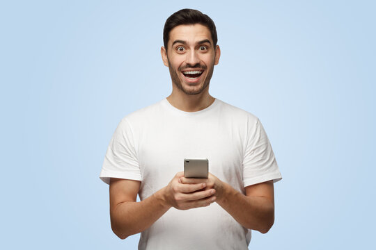 Young attractive laughing European man in white T-shirt holds cellphone in hands