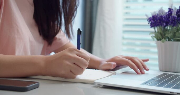 An woman Asian university college student uses a laptop computer on the table at his home.Female hands using a laptop and take notes on notepad.for studying online.Online education concept.