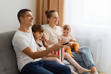 Side view portrait of family sitting on sofa with remote control and watching tv, enjoying interesting film or cartoon, being extremely happy being together at home.