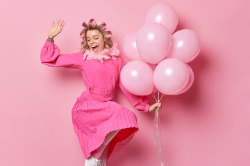 Obraz na płótnie Canvas Happy cheerful young woman wears festive dress dances against pink background with bunch of balloons applies hair rollers for making hairstyle has positive expression. Special occasion concept