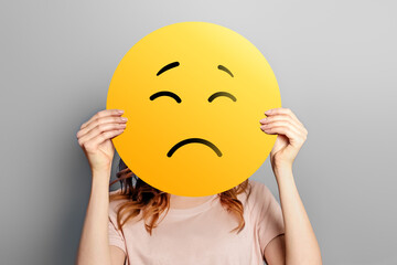Sad emoji. Girl holds a yellow emoticon with sad face isolated on a gray background. unhappy emoji