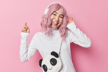 Carefree joyful Asian woman with pink hair catches every bit of song listens favorite music via headphones wears white turtleneck carries bag poses against rosy studio background. Good tunes in ears