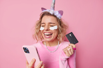 Bank clients. Positive beautiful woman holds credit card and smartphone laughs joyfully glad to win money undergoes beauty procedures wears unicorn headband dress and necklace isolated on pink © WHstudio Leushin N