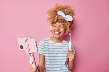 Glad young woman with curly hair wears headband and casual striped t shirt records video blog holds smartphone on selfie stick gives recommendations how to care about teeth holds toothbrush.