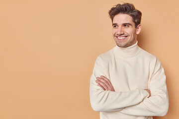 Studio shot of smiling confident young man keeps arms folded looks away notices something pleasant wears white turtleneck poses against beige background with mock up space for your promotion
