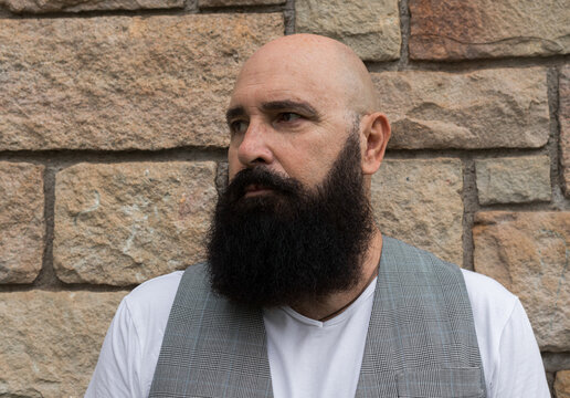 portrait of a shaved head mature man with nice black beard ahead of a brick wall and looking away
