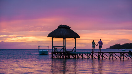 couple watching the sunset on a tropical beach with a wooden pier in the ocean, men and woman...
