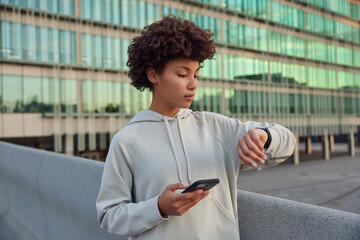 Waist up shot of active curly haired young woman uses phone application and smartwatch checks...