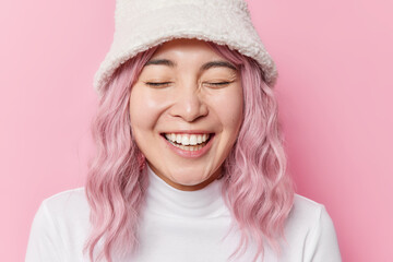 Cheerful overjoyed Asian woman with rosy hair laughs sincerely shows white teeth keeps eyes closed wears hat and poloneck isolated over pink background. People emotions and feelings concept.