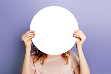Young woman holding round white poster covering her face isolated on lilac background in studio....