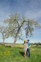 Fototapeta premium Loving couple near a blossoming tree on a background of spring blue sky. Girl with a wreath of dandelions on her head.