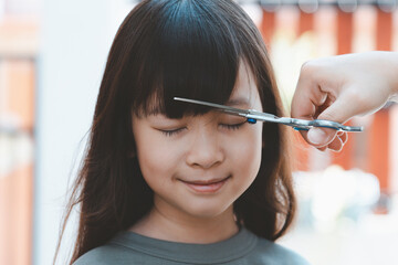 Women's hands to cut the front hair or bangs for a cute Asian girl at home. Mothers are happy to cut their children's hair. Hair care concept