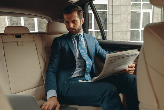 Elegant bearded businessman in classic clothes with newspaper in hand in back seat of car