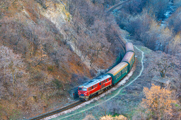 A beautiful red locomotive with an intercity passenger train rides along a mountain railway road. Aerial view.