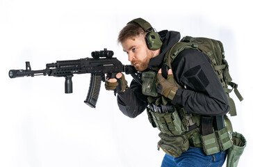 Fototapeta premium airsoft player in full gear with GG RK74 fire series guns. a man in headphones, a bulletproof vest, with a backpack and a belt, aims his machine gun to the side. profile. White background.