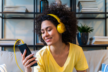Beautiful latino woman with curly hair at home listining music on mobile phone app with headphones