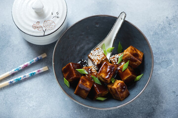 Bowl with pan fried tofu cubes in teriyaki sauce, high angle view on a light-blue stone background, studio shot