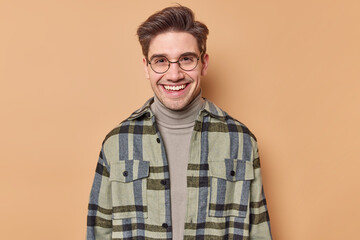 Portrait of handsome brunet European man smiles gladfully looks directly at camera wears round spectacles turtleneck and checkered shirt isolated over brown background. Happy emotions concept