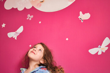 Young cute girl wearing pajamas looking up at a pink wall with decal stars, moon and fairies - obrazy, fototapety, plakaty
