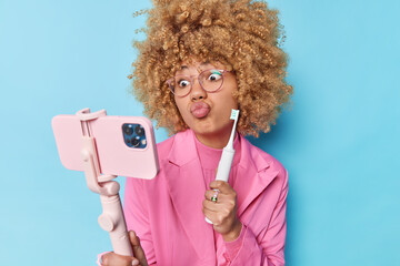 Funny blonde curly haired woman pouts lips makes grimace at smartphone camera holds electric...