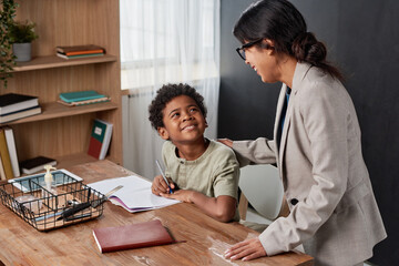 Hispanic tutor in jacket touching shoulder of black boy while supporting him during one-by-one lesson
