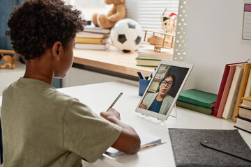 African American boy with curly hair sitting at table in nursery room and making notes while listening to online lecture