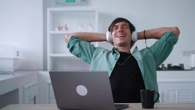Young man in wireless headphones is resting after working at a computer, enjoying good mood. Freelancer rejoices at a successfully completed job at a laptop while sitting at the kitchen table at home