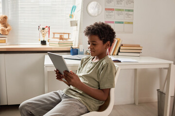 Smiling African American boy with curly hair sitting on chair and using tablet in own room