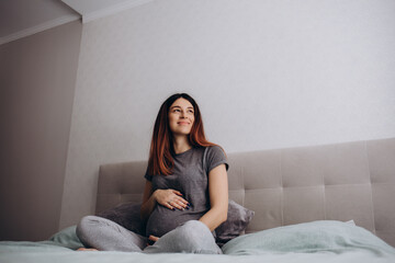 Obraz premium Attractive pregnant woman is sitting in bed and holding her belly. Last months of pregnancy.