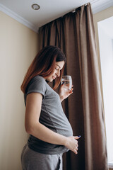 Hydration. Positive Pregnant Girl Drinking Water From Glass Standing Looking In Window Indoor.