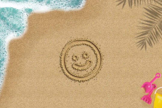 Smiley face drawn on sandy beach, travel and holiday concept, emoji idea, palm shadow and sea wave and toys composition, top view of sand beach