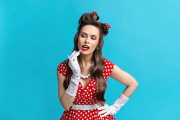 Beautiful young pinup woman in retro dress applying bright red lipstick over blue studio background