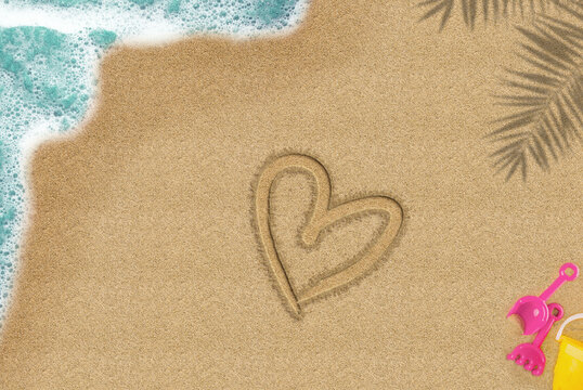 Heart drawn on sandy beach, trip and valentine holiday concept, top view of sand beach, summer times valentine idea, palm shadow and sea wave and toys composition