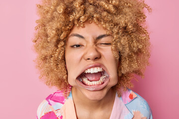Headshot of curly haired young woman uses mouth expander special tool for whitetning shows white...