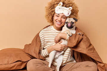 Cheerful young woman embraces her beloved pet poses in bed with pug dog covered with soft blanket smiles gladfully enjoys company of favorite domestic animal isolated over brown studio background.