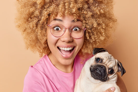 Portrait of happy woman poses with favorite pet laughs gladfully wears spectacles and pink casual t shirt glad to have new family member gets puf dog as present isolated over brown background