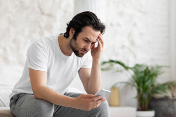 Upset young man using smartphone in bed