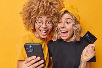 Happy amazed young women make online shopping use mobile phone and credit card download application to making payment use modern technologies dressed casually isolated over yellow background