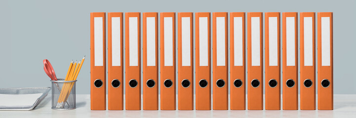 A row of mockup orange document binders or lever arch file with paperwork on an office shelf or desk. Paper tray and pencils. Business concept banner. Copy space