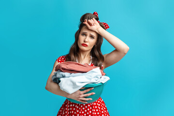 Tired young pinup woman in retro style dress holding clothes for washing or ironing, wiping...