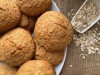Oatmeal cookies on a white plate. Oat flakes in a measuring spoon. Cookies on brown background top view.