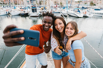 Happy young friends talking selfie with cellphone while drinking beer on a boat in summer vacation