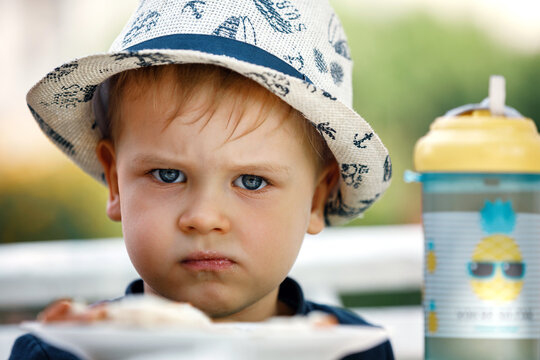Close up portrait of small angry caucasian boy in hat unwilling to eat food.