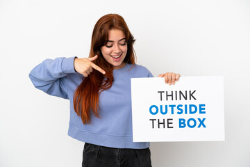 Young redhead woman isolated on white background holding a placard with text Think Outside The Box and  pointing it