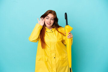 Redhead woman holding an umbrella isolated on blue background listening to something by putting hand on the ear