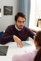 Young bearded tutor in sweater sitting at table with tablet and explaining difficult topic to elementary student girl