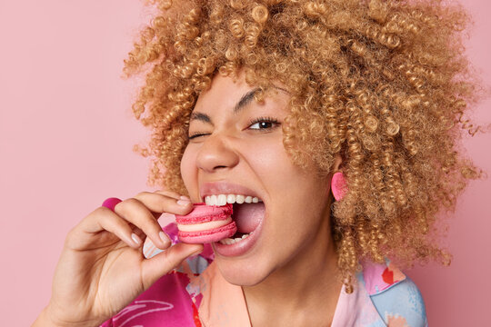 Close up shot of pretty curly haired woman eats delicious french macaroon enjoys dessert bakery sweet yummy food keeps mouth widely opened poses against pink background. Unhealthy eating concept