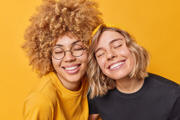 Portrait of happy young European women keep eyes closed from satisfaction smile toohily being in good mood dressed in casual t shirts express positive emotions isolated over yellow background